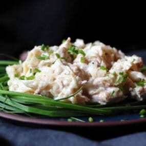 A plate of creamy chicken salad garnished with chopped chives and surrounded by whole chives on a dark background.