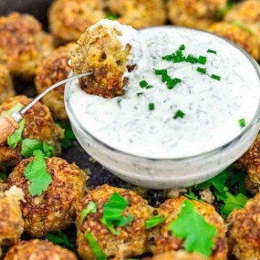 A plate of meatballs garnished with parsley surrounds a bowl of creamy white dipping sauce topped with chopped chives. A fork holds one meatball dipped in the sauce.
