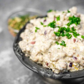 A bowl of creamy dip garnished with chopped green herbs, placed on a gray surface.