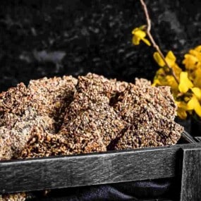 A wooden tray filled with sesame seed brittle sits next to a small metal container holding yellow flowers.