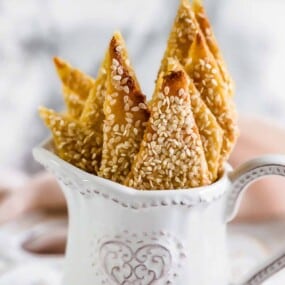 Several triangle-shaped sesame-covered pastries are arranged in a decorative white ceramic pitcher with a heart design.