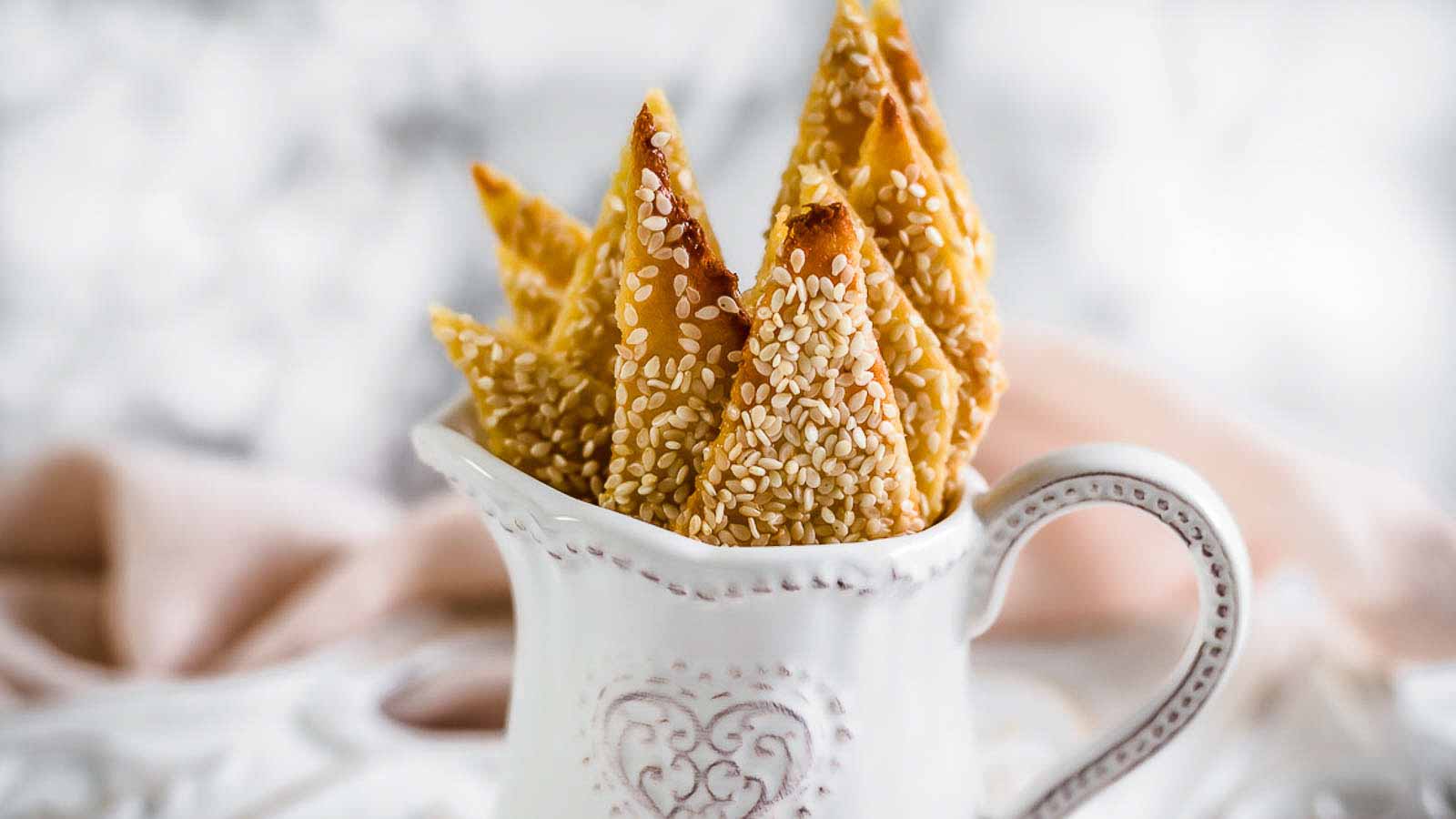 Several triangle-shaped sesame-covered pastries are arranged in a decorative white ceramic pitcher with a heart design.