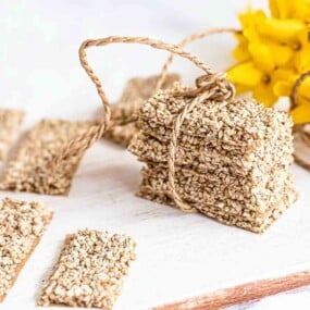 A stack of sesame seed bars tied with twine, surrounded by more sesame bars and a bunch of yellow flowers on a white surface.