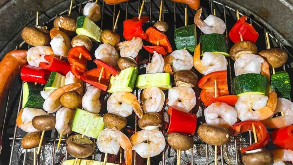 Skewers of shrimp, mushrooms, zucchini, and bell peppers grilling on a barbecue grill.
