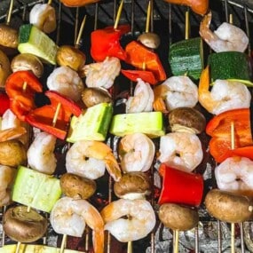Skewers of shrimp, mushrooms, zucchini, and bell peppers grilling on a barbecue grill.