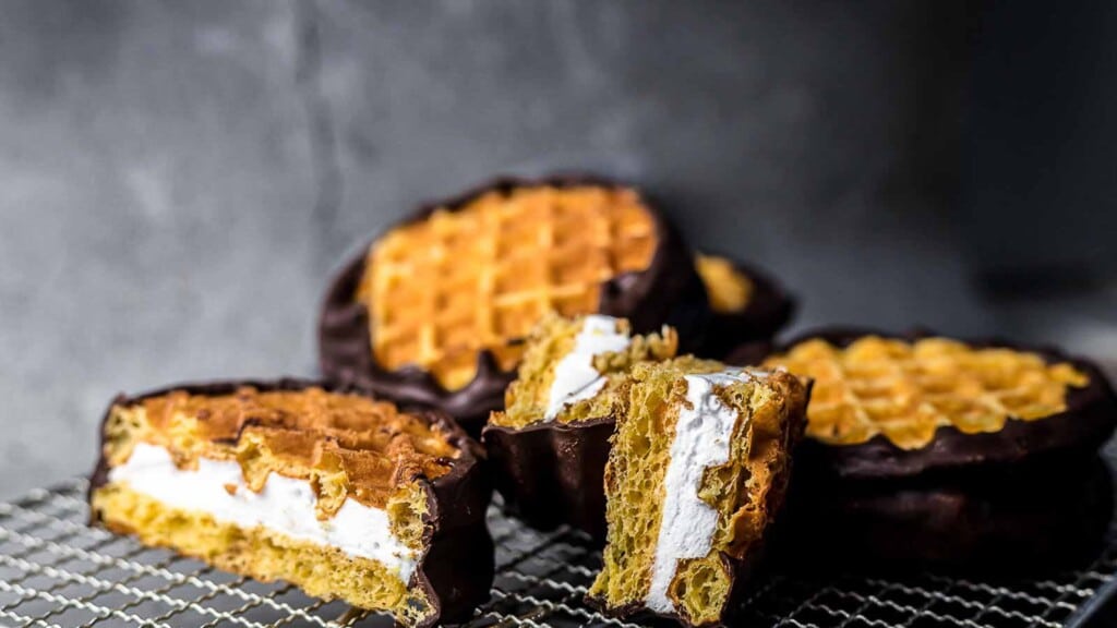 Waffle sandwiches filled with cream and partially dipped in chocolate are arranged on a wire cooling rack.
