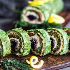 A sliced rolled spinach and salmon roulade on a wooden board, garnished with dill and lemon peel.