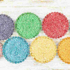Eight colorful beaded coasters in shades of purple, green, red, blue, yellow, and orange are arranged on a white wooden surface.