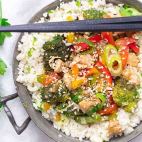 A bowl of mixed vegetables and chicken stir-fry sprinkled with sesame seeds over a bed of white rice, with chopsticks resting on the bowl and fresh parsley on the side.