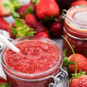 Two jars of strawberry jam, one open with a spoon inside, surrounded by fresh strawberries on a wooden surface.