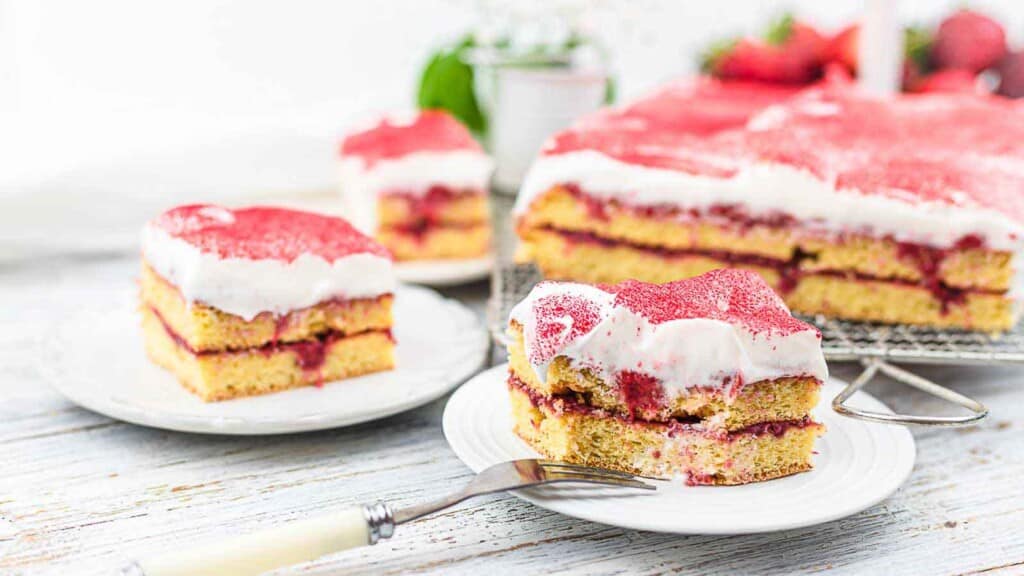 Two servings of layered cake with white frosting and strawberry filling are displayed on white plates, with a larger portion of the cake in the background on a wire rack.