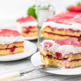 Two servings of layered cake with white frosting and strawberry filling are displayed on white plates, with a larger portion of the cake in the background on a wire rack.