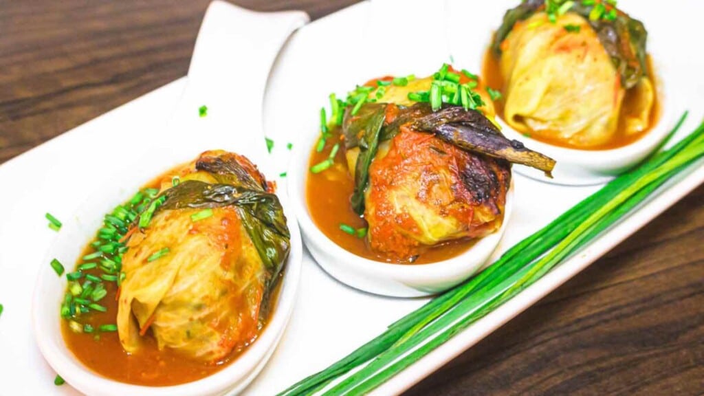 Three stuffed cabbage rolls served in white dishes, topped with green herbs and placed on a white rectangular plate with green garnish, on a wooden surface.