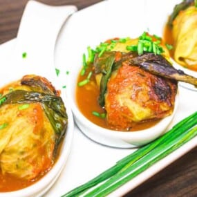 Three stuffed cabbage rolls served in white dishes, topped with green herbs and placed on a white rectangular plate with green garnish, on a wooden surface.