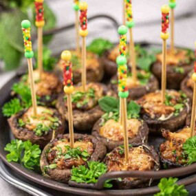 Stuffed mushrooms on a plate, each with a decorated skewer, garnished with fresh parsley and served on a metal tray.