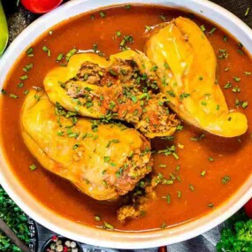 A white dish filled with stuffed yellow bell peppers in a tomato-based sauce, garnished with fresh herbs. Various fresh vegetables and herbs are arranged around the dish.