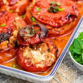 Baked stuffed tomatoes with a meat and rice filling in a baking dish, accompanied by a small bowl of fresh herbs.