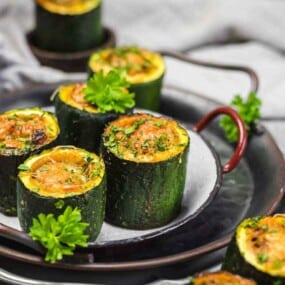 Stuffed zucchini cups filled with a savory mixture, garnished with fresh herbs, served on a gray plate with a sprig of parsley.