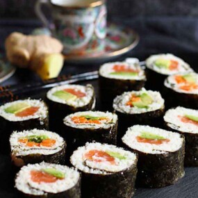 A plate features neatly arranged sushi rolls filled with vegetables and seafood, accompanied by pickled ginger, and a small dish of wasabi. Two decorated teacups and saucers are in the background.