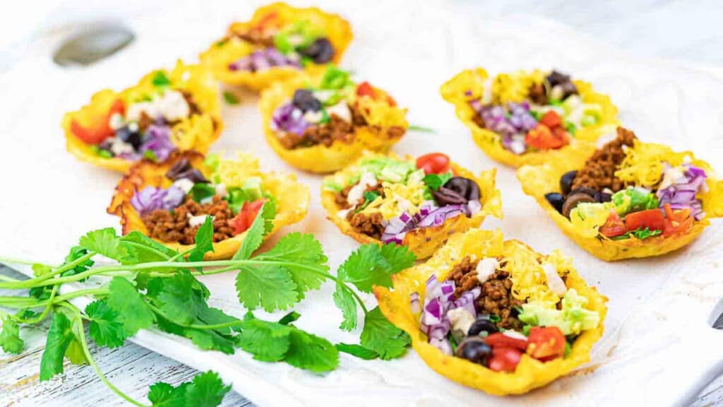 Mini taco bites filled with ground meat, tomatoes, shredded lettuce, cheese, olives, and purple cabbage, served on a white platter with fresh cilantro garnish.