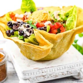 A taco salad served in an edible shell bowl. The salad contains lettuce, tomatoes, shredded cheese, black olives, and dressing, and is accompanied by a jar of seasoning on the side.