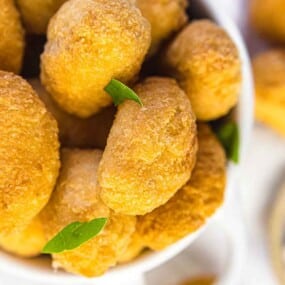 Close-up of a bowl filled with golden-brown, crispy croquettes garnished with small green leaves. Additional croquettes and a dip are partially visible in the background.