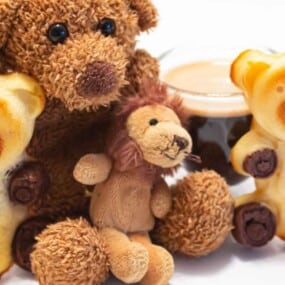 Two teddy bear-shaped pastries and two stuffed animals, a bear and a lion, are placed on a white surface. A glass of coffee is in the background.