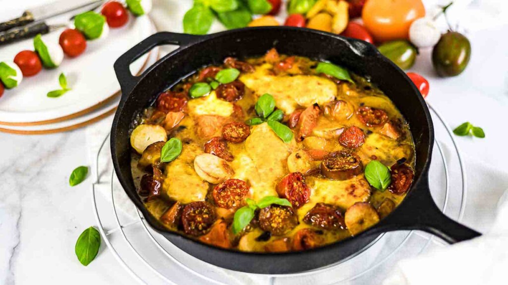 A black cast-iron skillet filled with a cooked dish of chicken, tomatoes, sausage, and basil, displayed on a white surface with various ingredients, including tomatoes and greens, in the background.