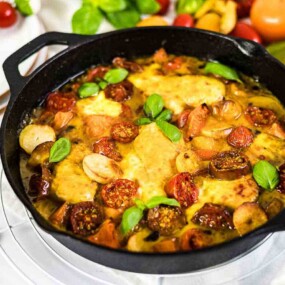 A black cast-iron skillet filled with a cooked dish of chicken, tomatoes, sausage, and basil, displayed on a white surface with various ingredients, including tomatoes and greens, in the background.