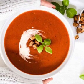 A bowl of tomato soup garnished with croutons and a basil leaf, held by hands in white sleeves. Another container of croutons and a few basil leaves are also on the white surface.