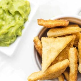 A white bowl of golden-brown tortilla chips is next to a small white dish of guacamole on a white background.