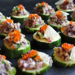 Sliced cucumber rounds topped with a mixture of finely chopped vegetables and herbs, decorated with small orange roe and garnished with lemon wedges, arranged on a dark serving platter.