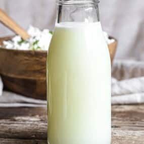 A glass bottle of milk is placed on a rustic wooden table with a wooden bowl containing cottage cheese in the background.