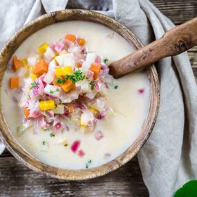 A bowl of creamy soup with diced vegetables, including carrots, corn, and onions, garnished with herbs, with a wooden spoon resting in it, placed on a wooden table.