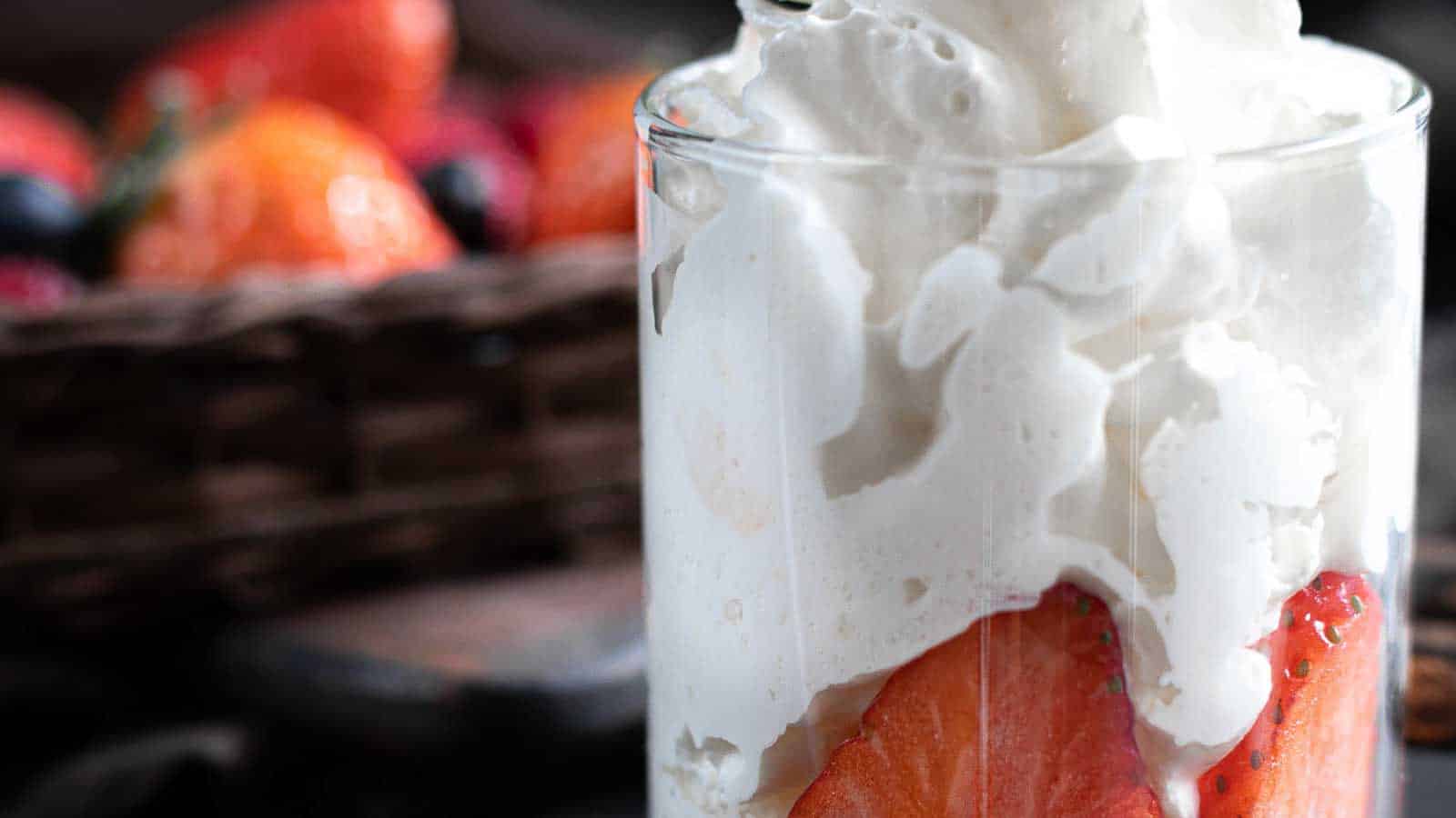 Close-up of a glass filled with layered strawberries and whipped cream, with a basket of mixed berries in the blurred background.