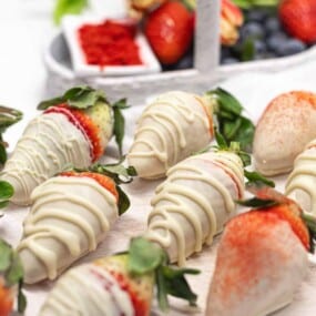 A selection of strawberries coated in white chocolate and drizzled with more white chocolate are arranged on a white surface. Additional strawberries and some blueberries are visible in the background.