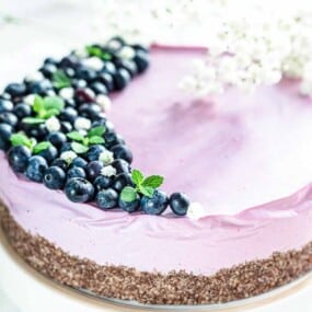 A round purple cake topped with blueberries and small green leaves on one side, displayed on a white surface with white flowers in the background.