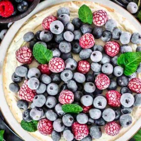 A cheesecake topped with a mix of frozen blueberries and raspberries, garnished with mint leaves. Bowls of extra berries and mint leaves are nearby.