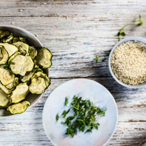 A wooden table with bowls of sliced zucchini chips, chopped herbs, and a small bowl of sesame seeds. Fresh sprigs of herbs are scattered on the table.