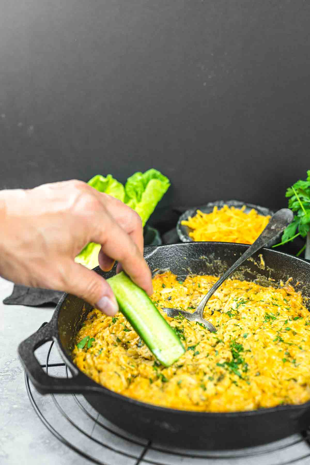 A hand dips a cucumber stick into a cast iron skillet filled with a cheesy dip. Lettuce leaves, a bowl of grated cheese, and fresh herbs are in the background.