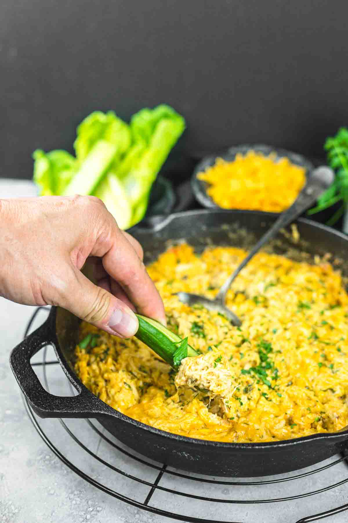 A hand dipping a piece of cucumber into a skillet filled with a cheesy casserole. Lettuce and a bowl of shredded cheese are in the background.