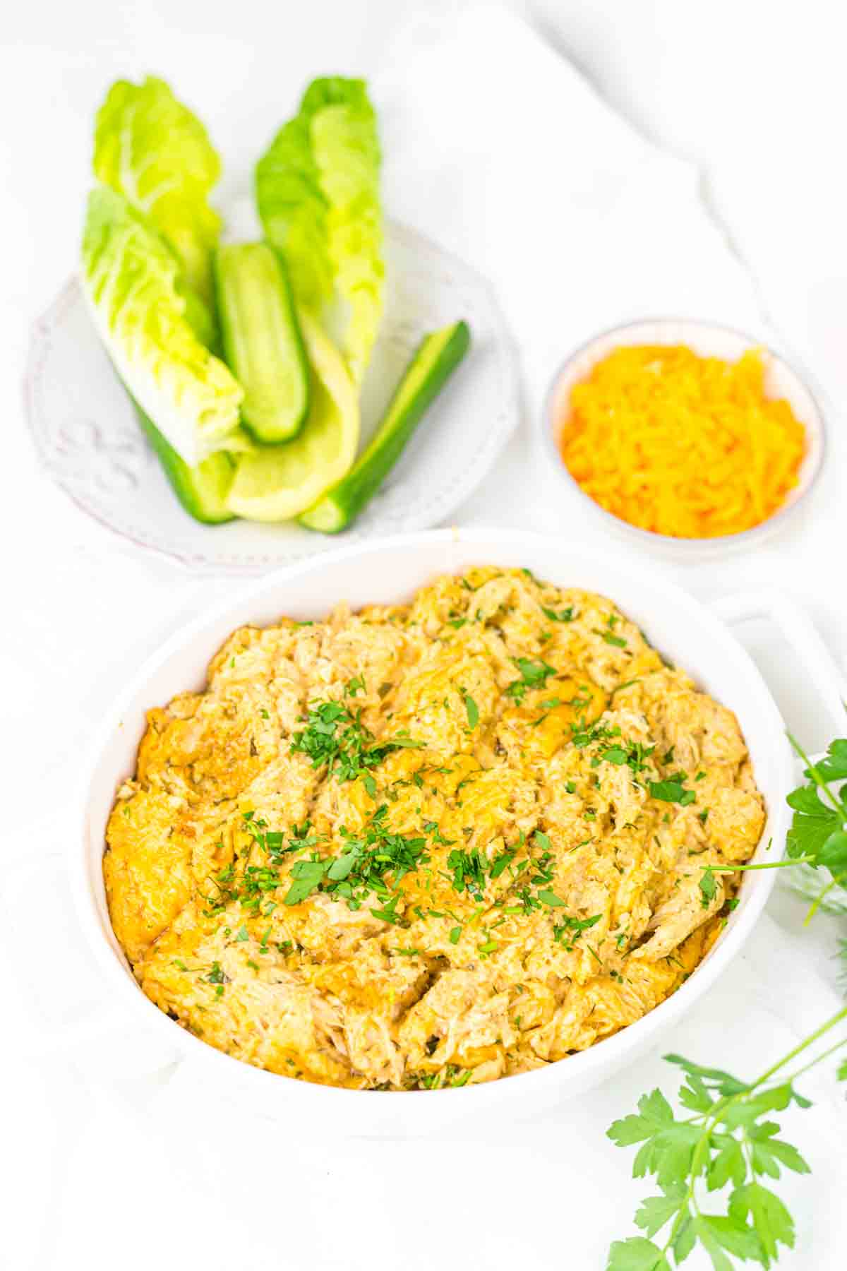 A casserole dish filled with a creamy chicken mixture topped with melted cheese garnished with parsley, a plate of lettuce leaves and cucumber, and a small bowl of grated cheese on the side.