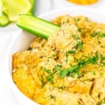 A close-up of a white bowl filled with a creamy, cheesy dish topped with chopped herbs. A slice of cucumber is dipped into the mixture.