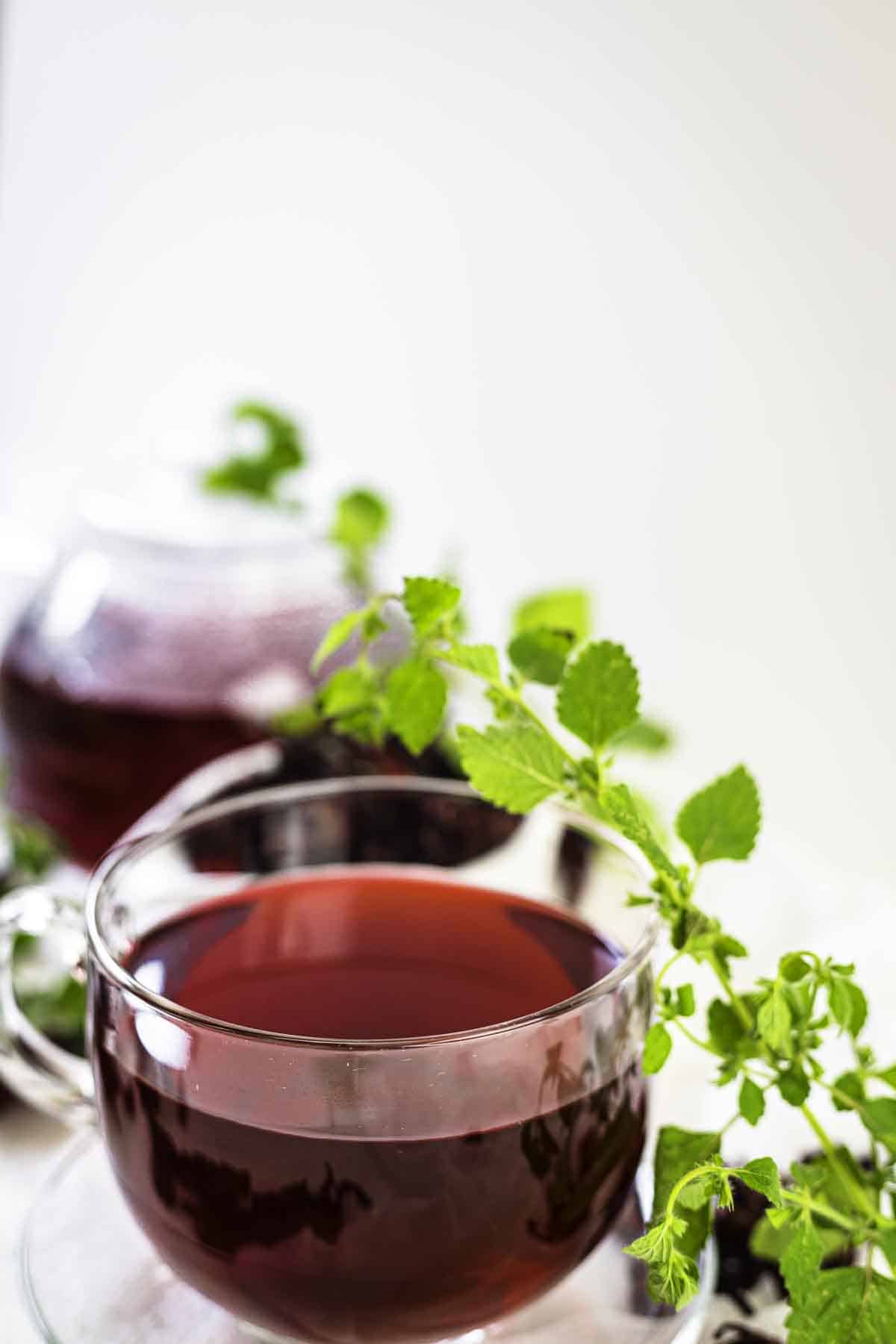 A clear glass cup filled with dark red tea sits on a white surface, accompanied by a green leafy sprig. Another glass teapot with more tea is visible in the background.