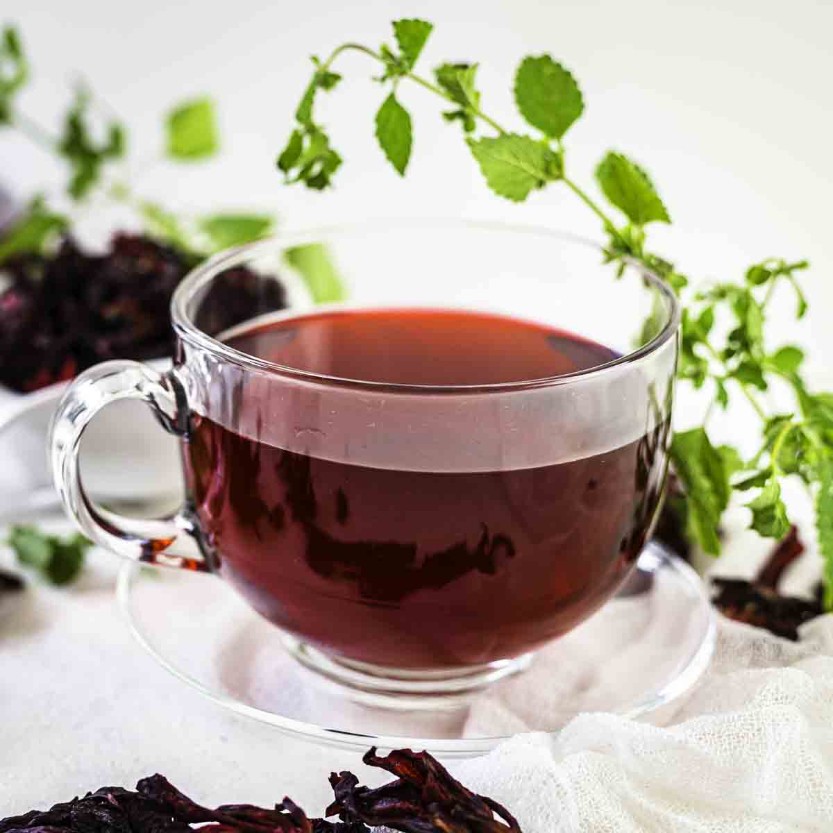 A glass cup filled with hibiscus red tea, on a saucer. Fresh green herbs and dried hibiscus flowers surround the cup. The background is white.