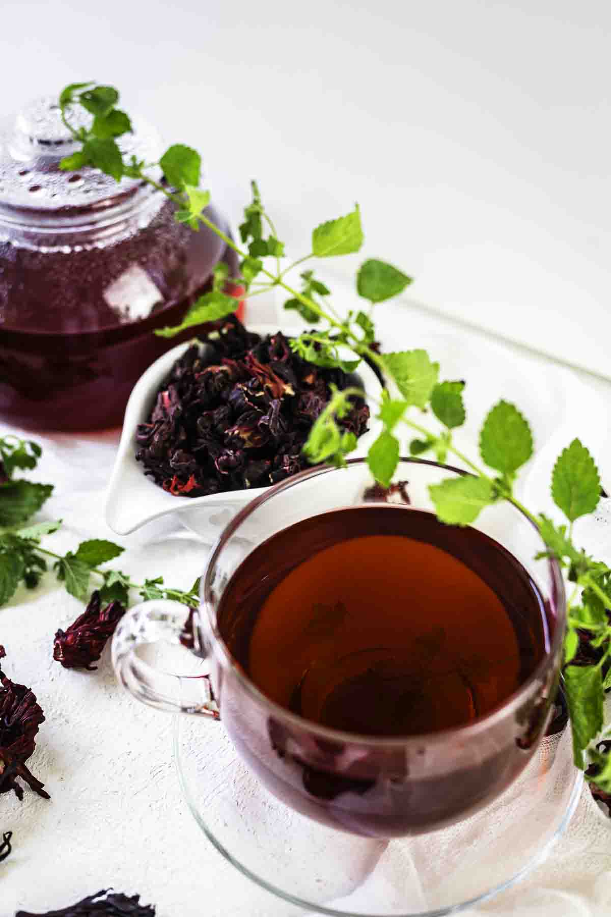 A glass cup filled with hibiscus tea, a small white bowl containing dried hibiscus flowers, and a glass teapot, all surrounded by fresh green leaves.