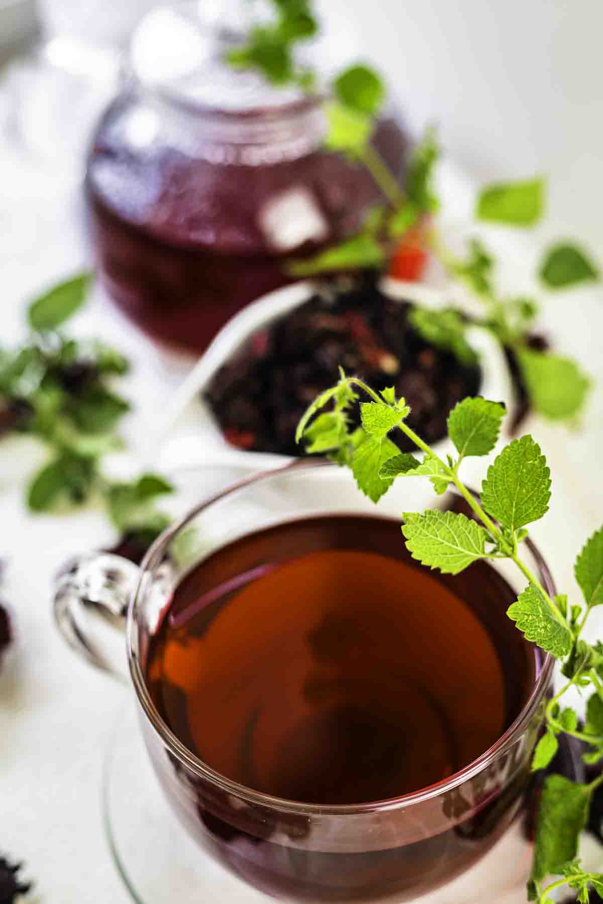 A clear glass cup of herbal tea garnished with fresh mint leaves beside a teapot and a bowl of tea leaves on a white surface.