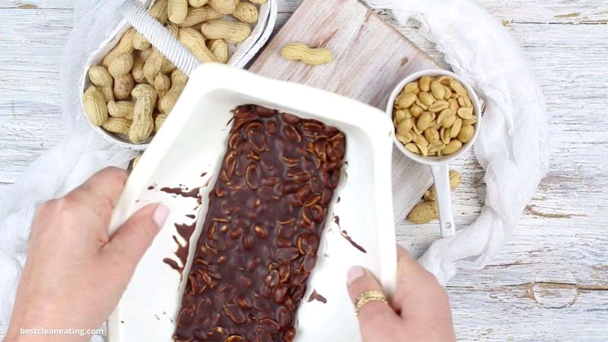 Hands holding a tray of chocolate-covered peanuts, with a bowl of peanuts, a bowl of shelled peanuts, and a few loose peanuts on a wooden surface.