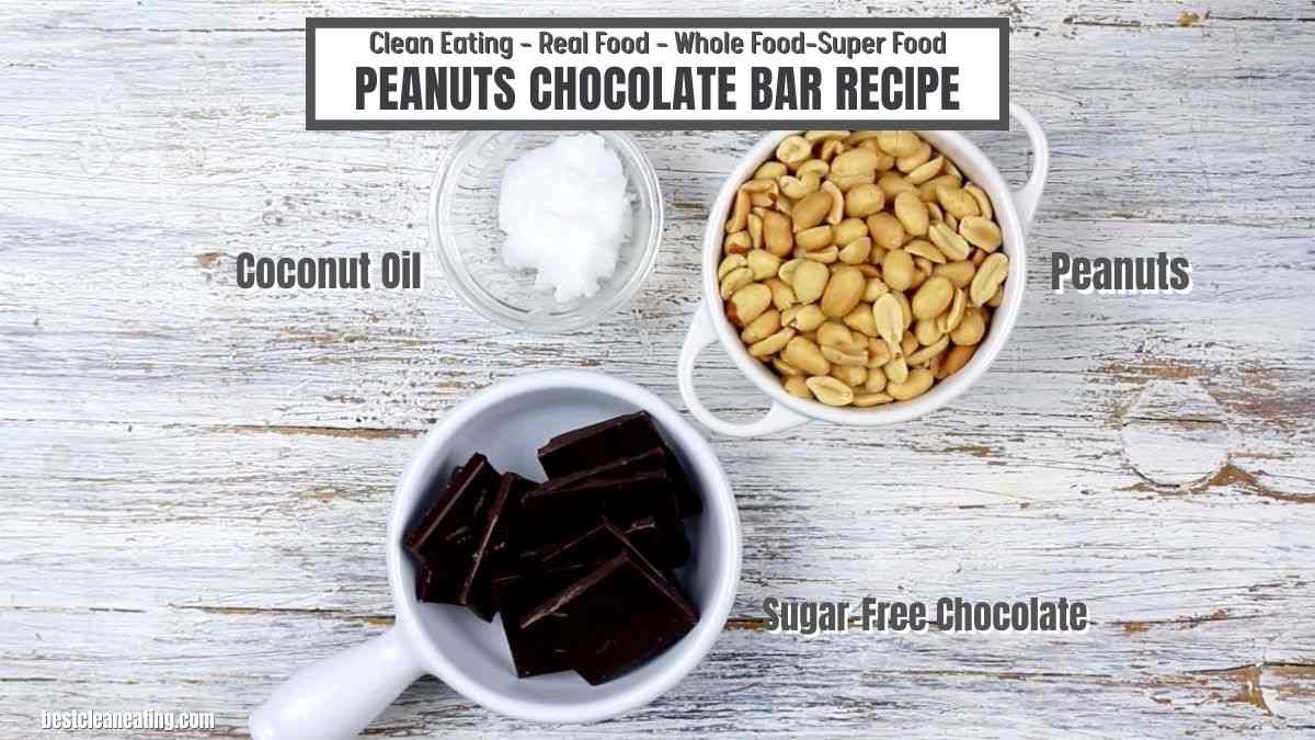 Top-down view of ingredients for a peanuts chocolate bar recipe: a bowl of peanuts, a bowl of coconut oil, and a bowl of sugar-free chocolate, displayed on a rustic wooden surface.