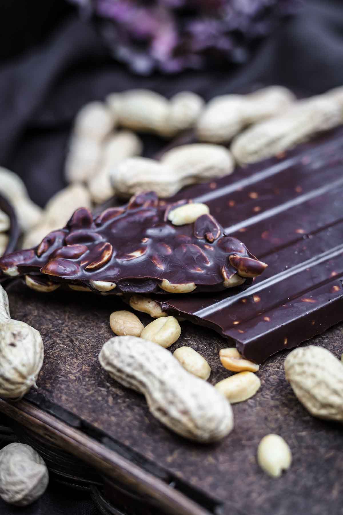 A dark chocolate bar with a piece containing embedded peanuts, surrounded by whole and shelled peanuts, rests on a wooden surface.